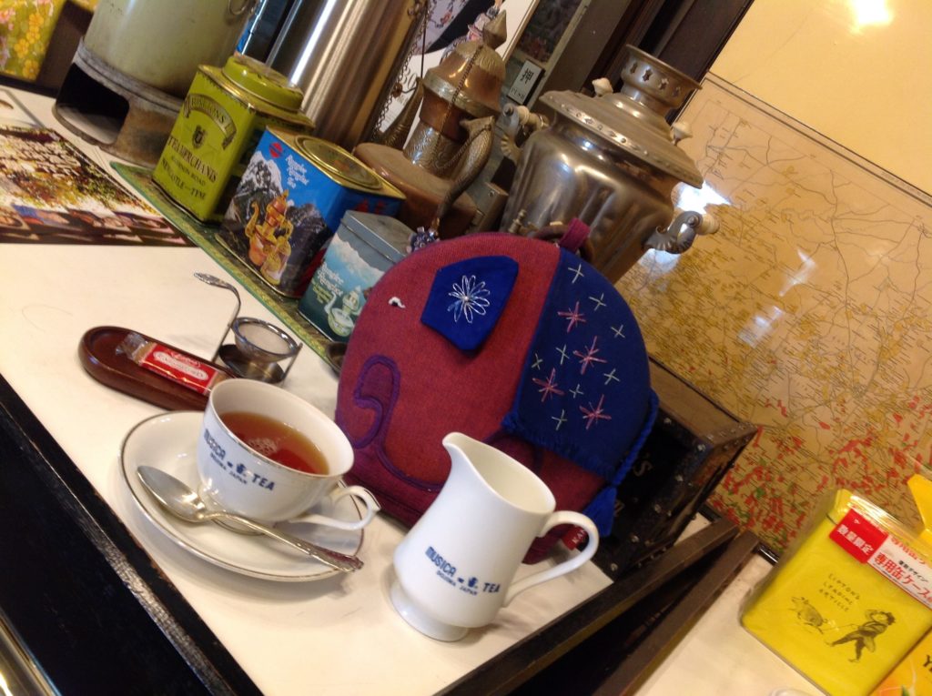 サモワールの前で紅茶。ティーコージーが被せられたポット、ティーカップの中に紅茶、ミルクジャグ、ロータスのカラメルビスケット一枚入り、回転茶こし。