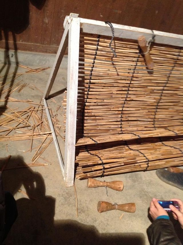 [12]編むの終了。重しはこうなってたんですね。 白いひもで黒い縄を括っていたらしい。 木に溝が入っているから、縄の位置がズレないんです。また、地べたの左側に目をやると、長くて切ったヨシの切れ端がたくさん落ちてます。